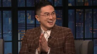 Bowen Yang on Late Night with Seth Meyers holding his hands in a prayer like position and smiling to his right.