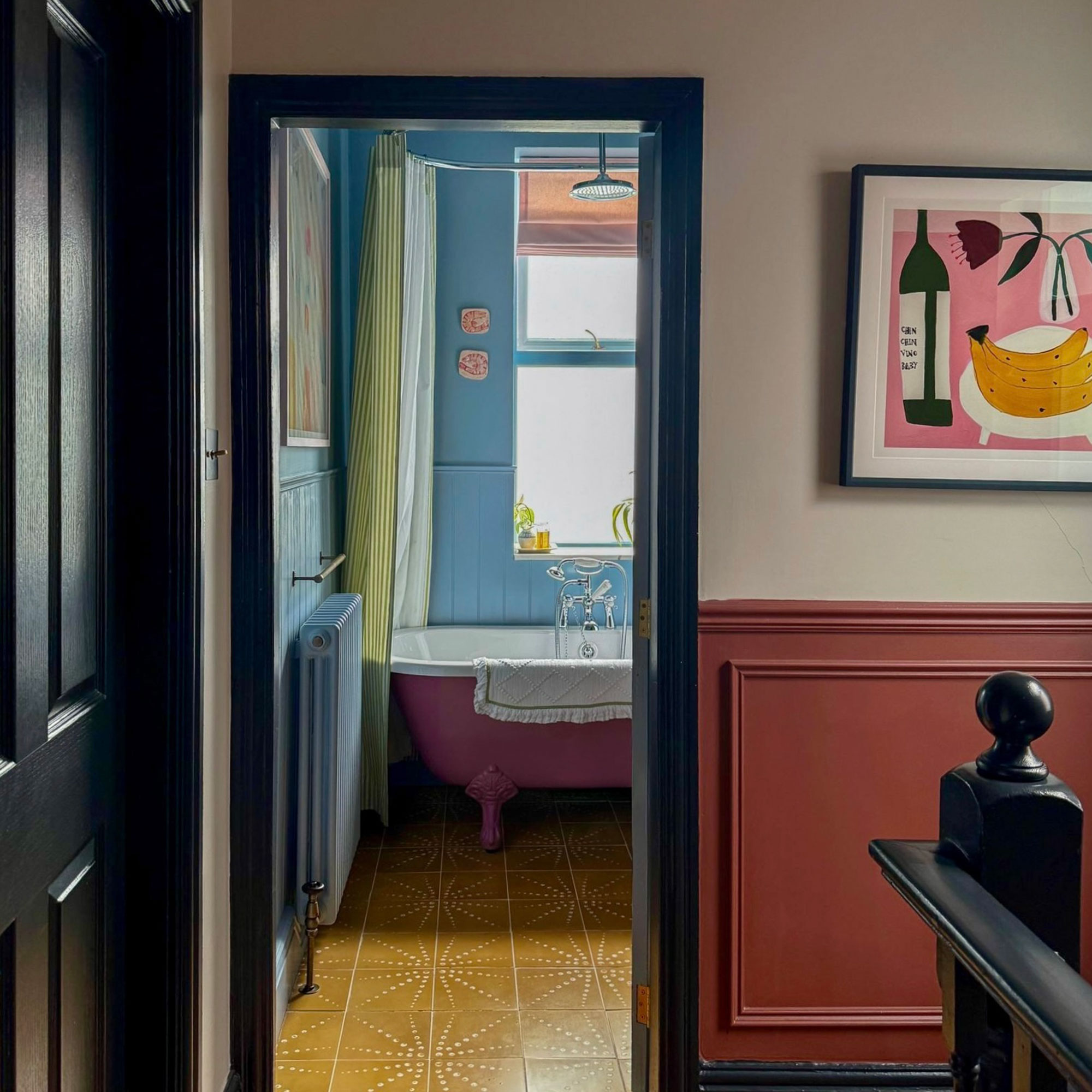 Upstairs landing with half neutral walls, half terracotta painted panelling, leading through to bathroom with blue walls, yellow floor tiles and pink freestanding bath