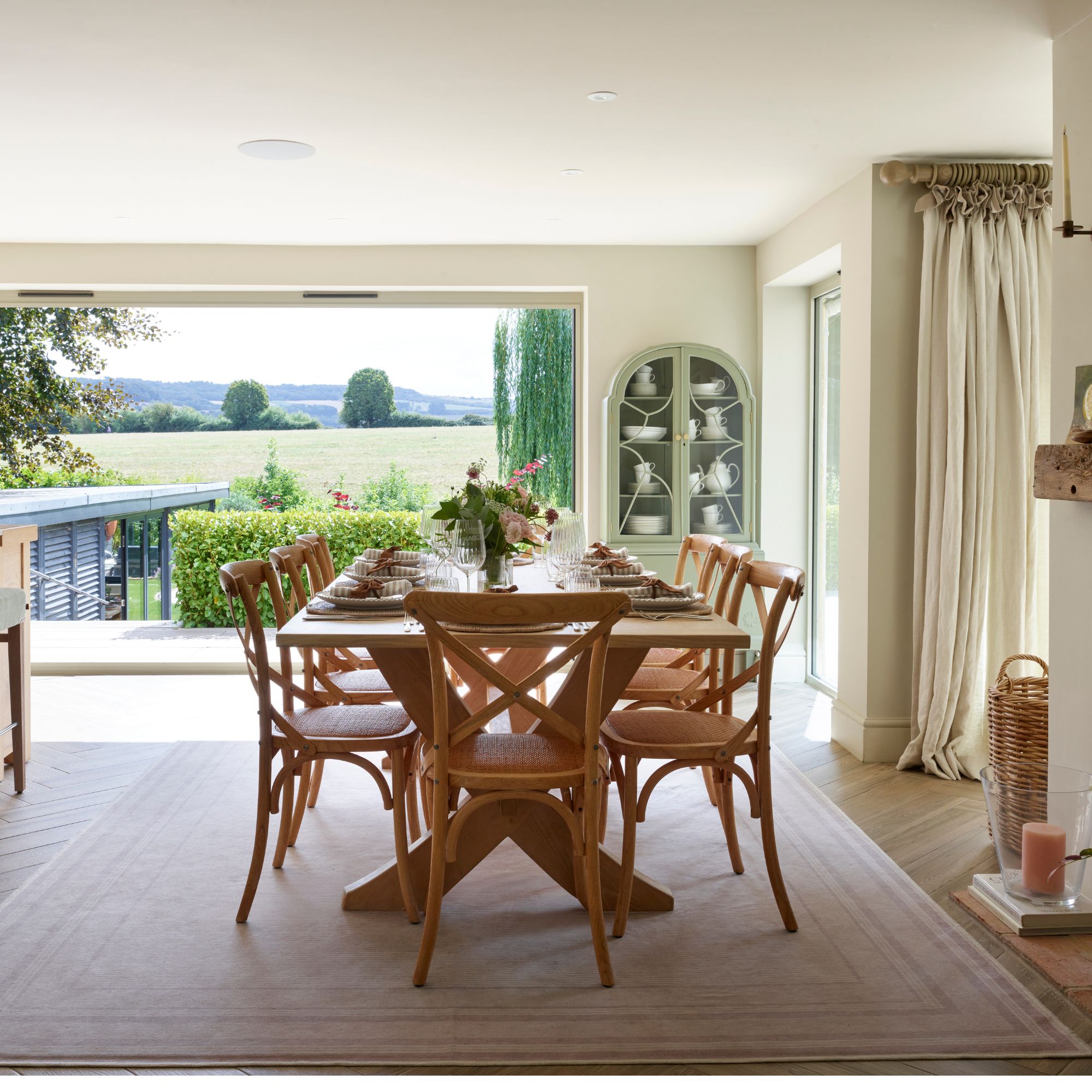 Dining room with wooden table and chairs and pale pink rug, green arched cabinet and large bifold doors opening to country views