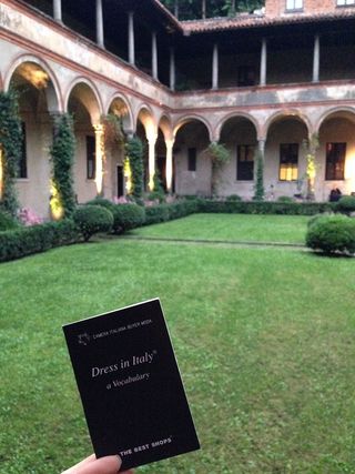 "Dress in Italy" book held by someone in a courtyard with grass and many green plants