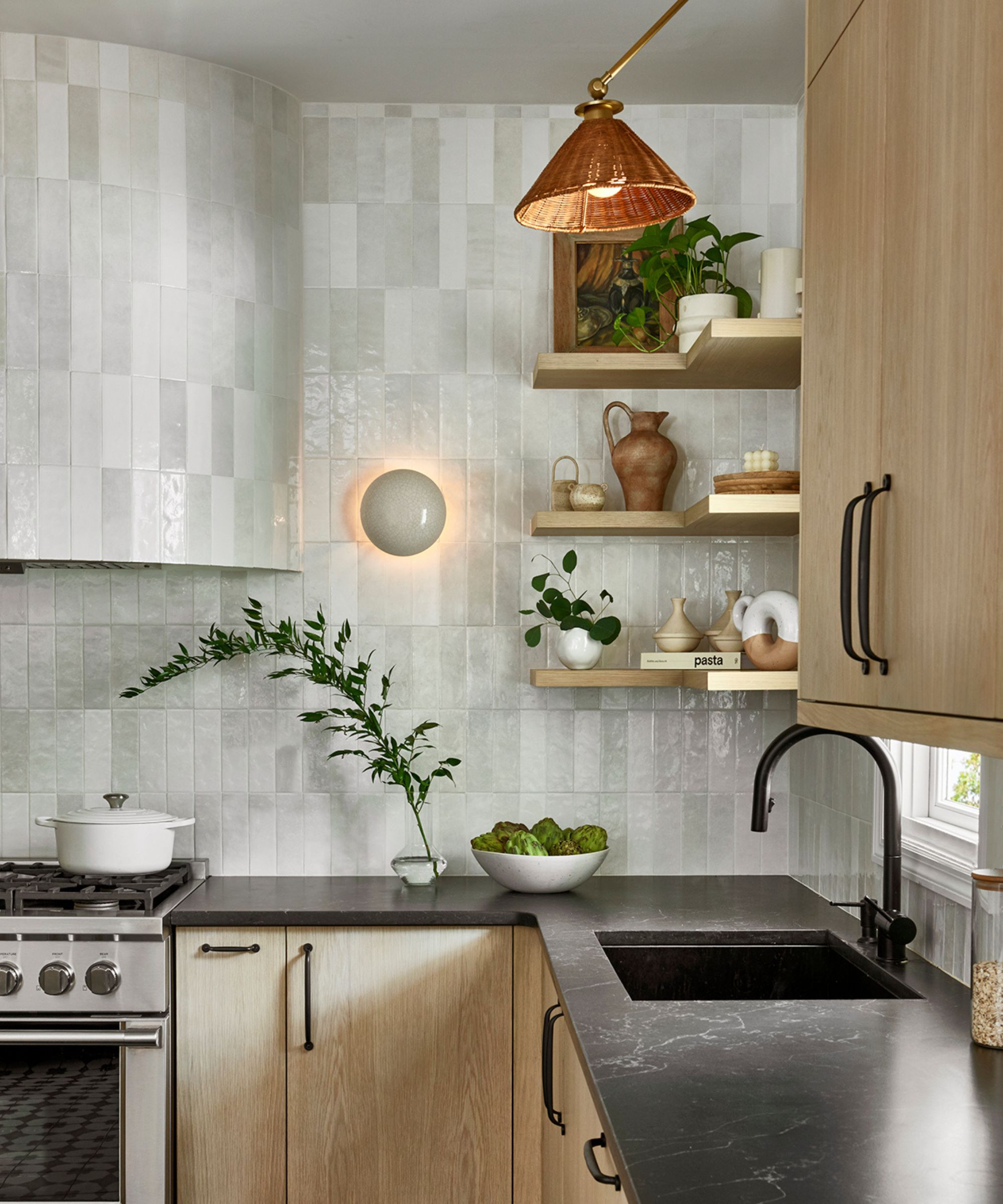 A wooden kitchen with white zellige backsplash tiles, black countertops, wooden shelving, and a rattan light shade