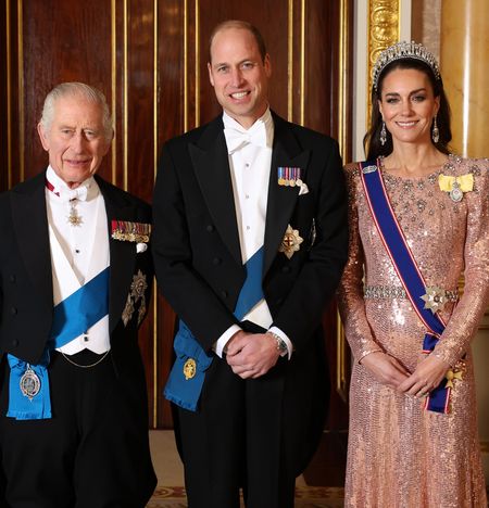 Princess Kate wearing a pink sequined gown and a tiara standing next to Prince William and The King wearing tuxedos