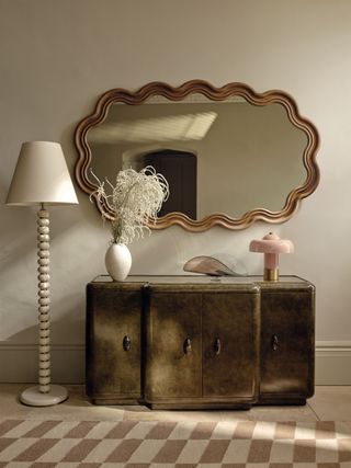 Entryway shot of a green-dyed cherry wood console cabinet. On the cabinet is a light pink mushroom lamp, a wavy glass dish, and a white vase with white stems in it. Above the cabinet hangs a horizontal wavy mirror. There is a white bobbin floor lamp and a dusty pink and off-white checkerboard rug.