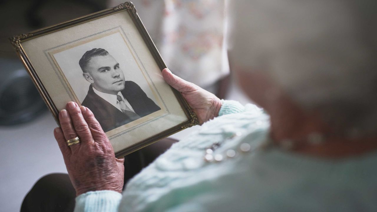 A woman with grey hair wearing a wedding ring holds a black-and-white photo of a man. 