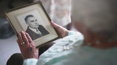 A woman with grey hair wearing a wedding ring holds a black-and-white photo of a man. 