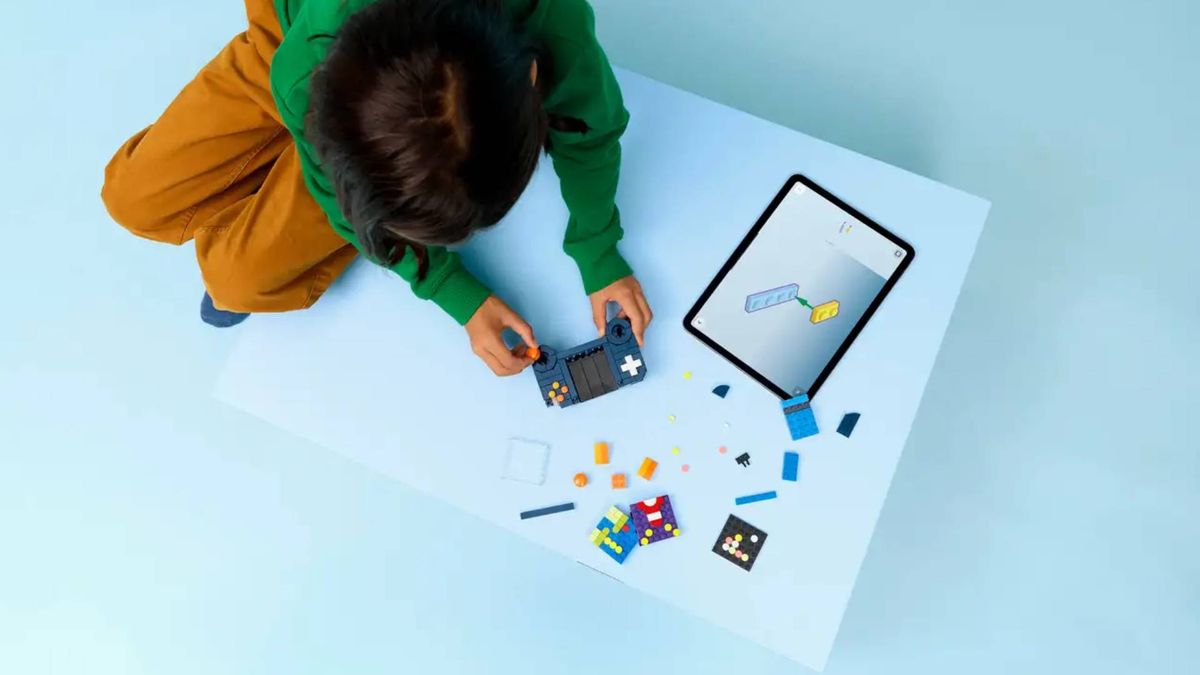 Kid with orange trousers and green jumper building Lego 3 in 1 Retro Game Console using tablet for instructions to make handheld.