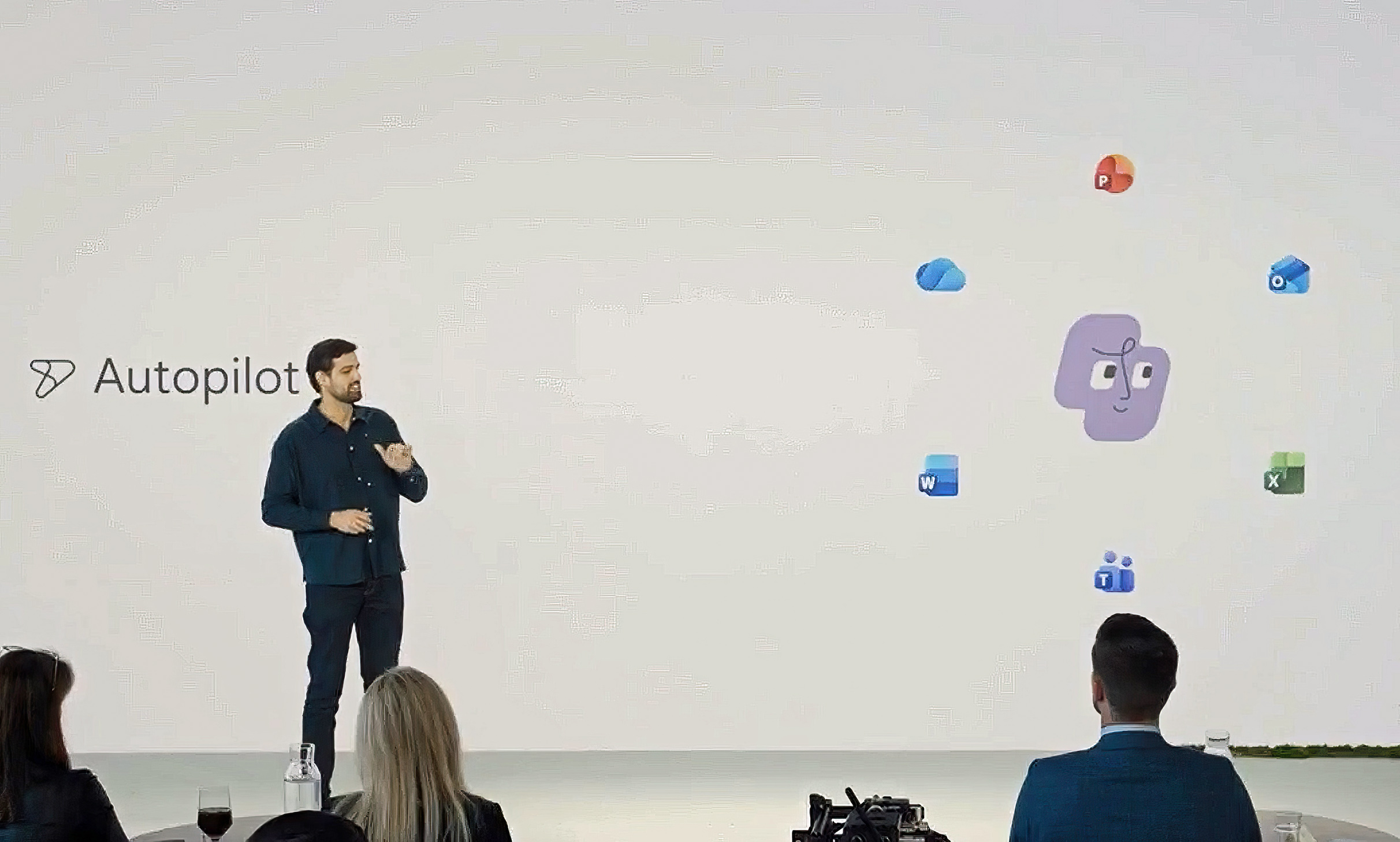 Jacob Andreou is on stage in front of a screen displaying "Autopilot" and Microsoft 365 app icons surrounding a central icon, with an audience seated at tables in the foreground.