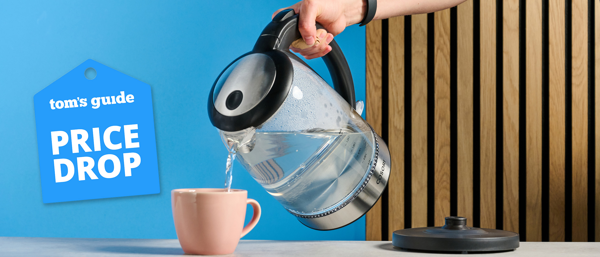The Cosori Electric Glass Kettle filled with water next to a pink mug photographed in front of a blue blue background with a price drop tag.