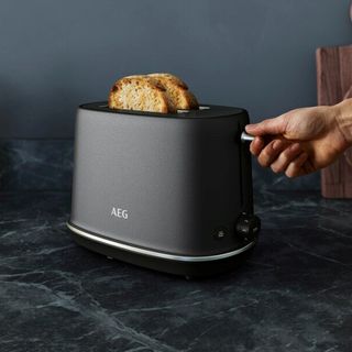 A black AEG toaster with toast inside it being popped up by a hand, on a dark black marble worktop