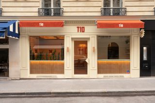 The facade of a modernly furnished restaurant features the sign "TIO" in pastel red on top of the main entrance, while two red curtains flag it in white. The interiors of the restaurant are warmly lit and intimate.