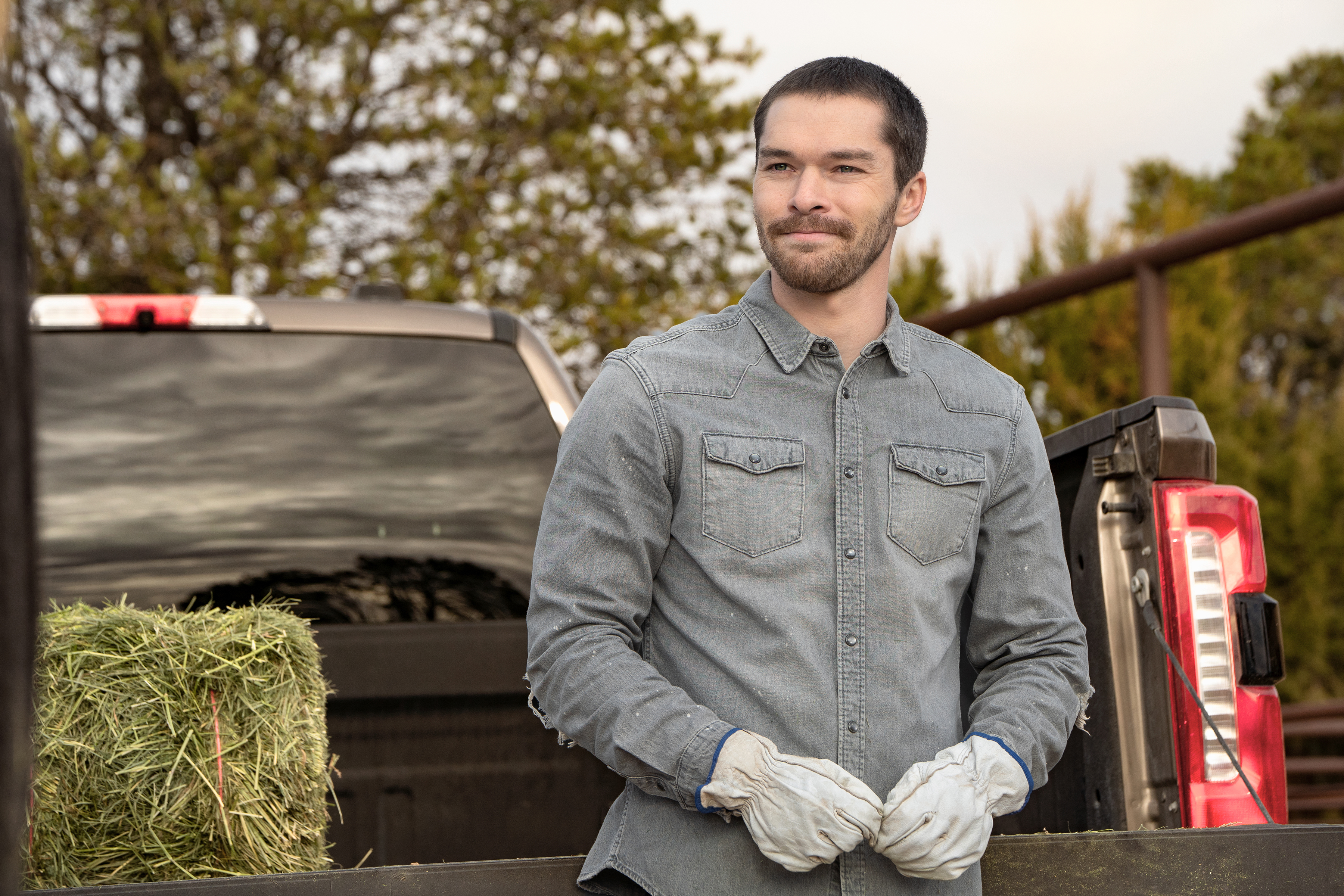 Casey W. Johnson as Kit leans on the bed of a pickup truck loaded with a bale of grass and a ladder, in Episode 206 of Netflix's 'Ransom Canyon' Season 2.
