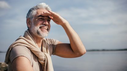 An older man shields his eyes as he looks into the distance next to a lake.