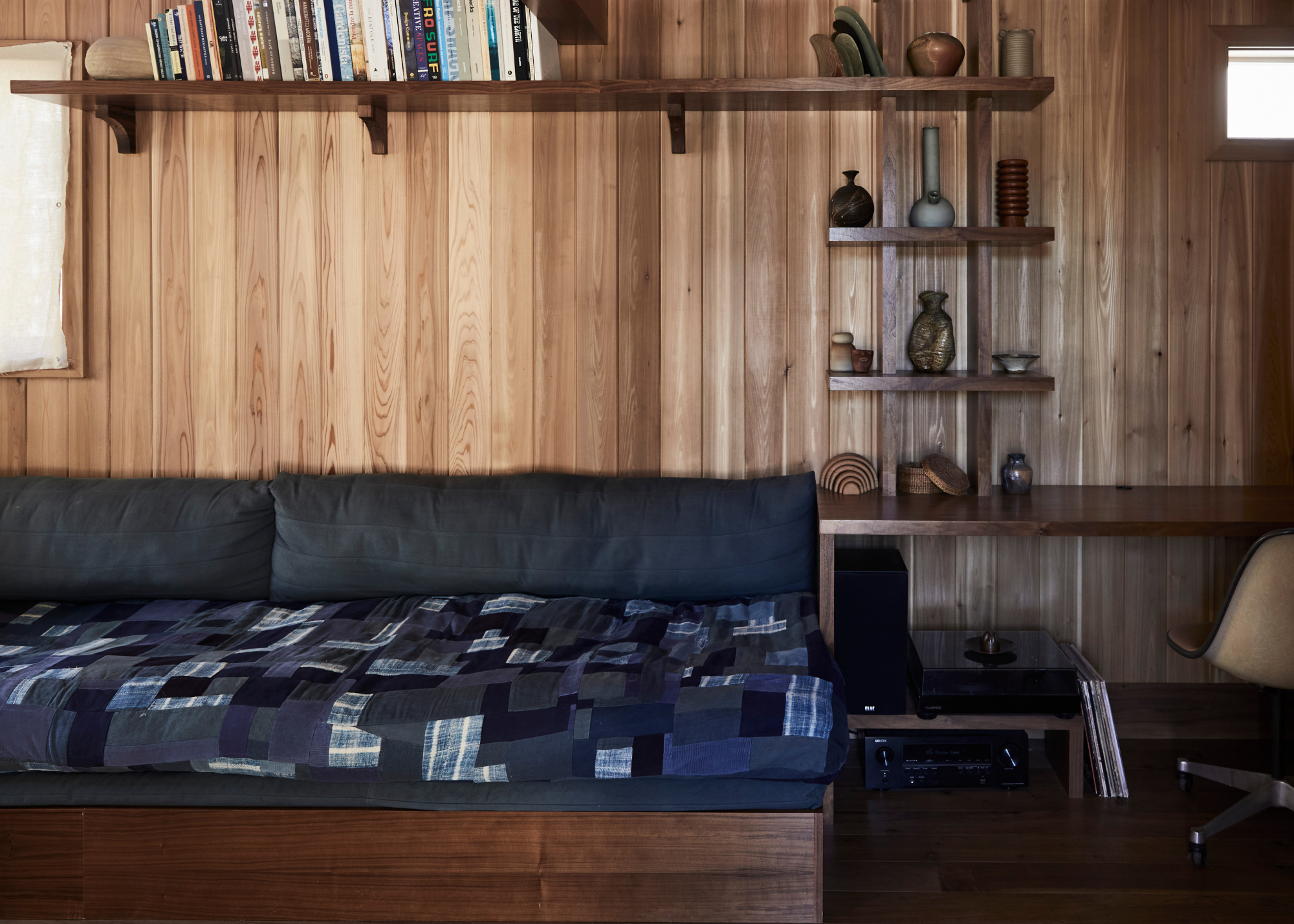 A room with a navy sofa bed, a patterned quilt, a vinyl stack, shelves of ceramics, and a swivel chair