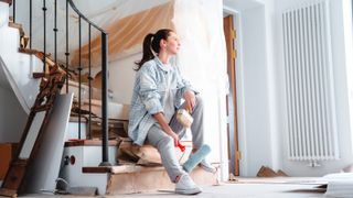 woman sat on staircase with paint roller in hand mid hallway renovation