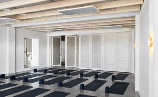 Interior view of a yoga studio at Studio Warrior One featuring white walls, light wood ceiling beams, white folding doors, wall lights and multiple black yoga mats laid out on the floor