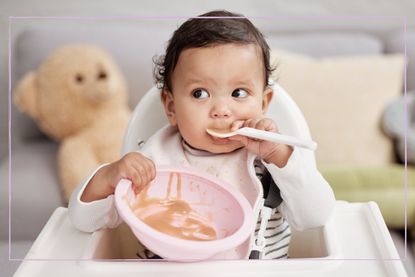 A baby with a sponnin their mouth holding onto a bowl