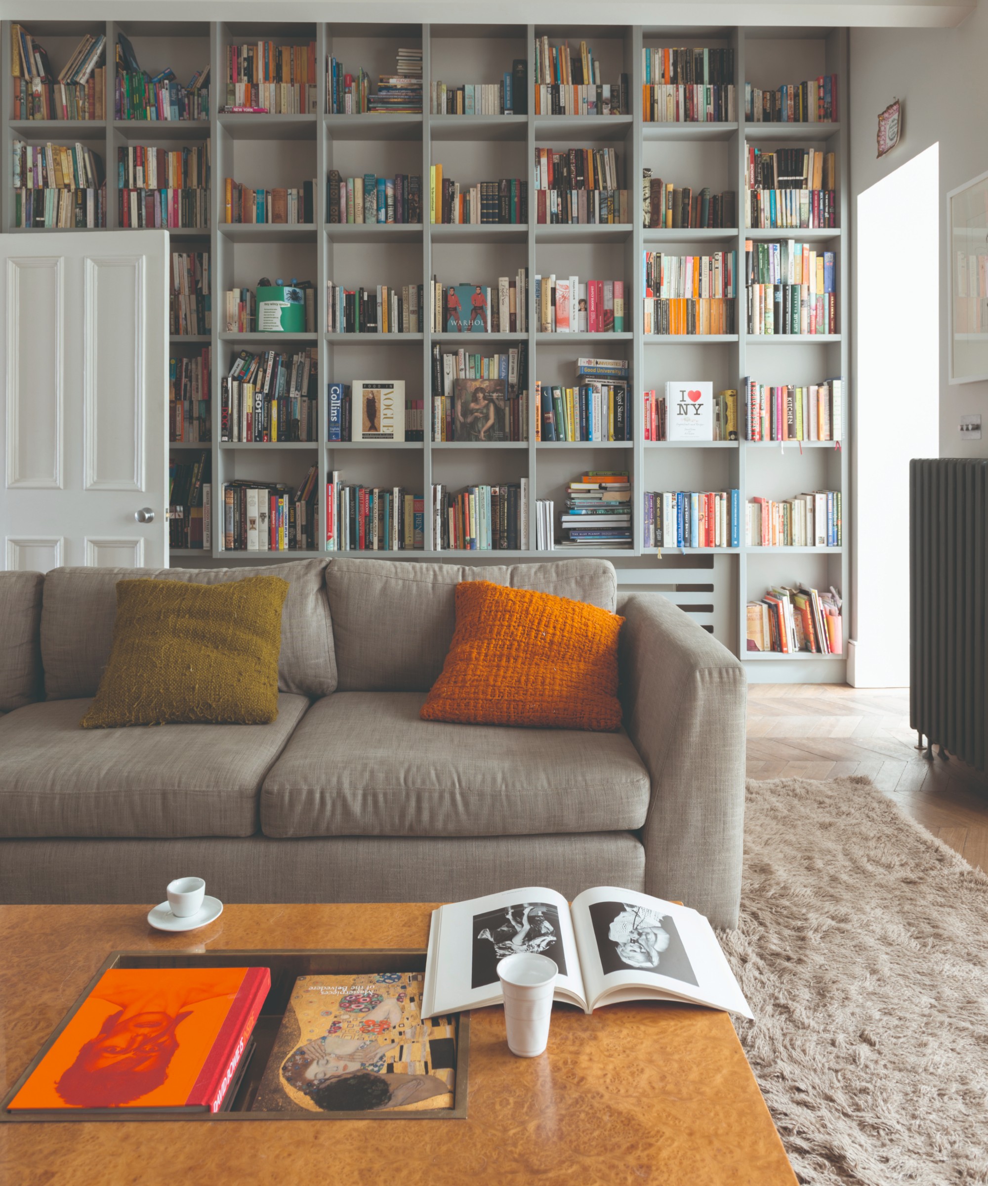 A grey living room with a grey sofa, orange accessories and a wall covered with a library