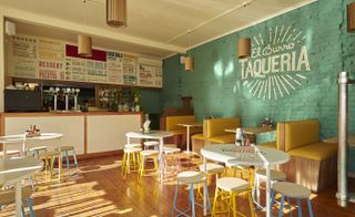 A view of the restaurant's counter with the menu displayed above in multiple artistic fonts and colours. White tables with condiments setup and colourful stools. Yellow leather and wood sofas with tables