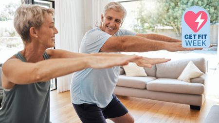 Older male and female smiling at each other performing a squat in the living room of a home