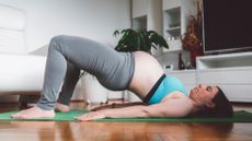 A pregnant woman performs glute bridge on an exercise mat. She is on her back, with her shoulders and feet against the floor, knees bent and butt elevated in the air. Behind her we see a TV, shelving and a leafy plant.