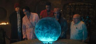 From left to right: Father Kent (Owen Wilson), Gabbie (Rosario Dawson), Ben Matthias (LaKeith Stanfield), Harriet (Tiffany Haddish) and Bruce Davis (Danny DeVito) stand around a table with a crystal ball on it in Haunted Mansion. 