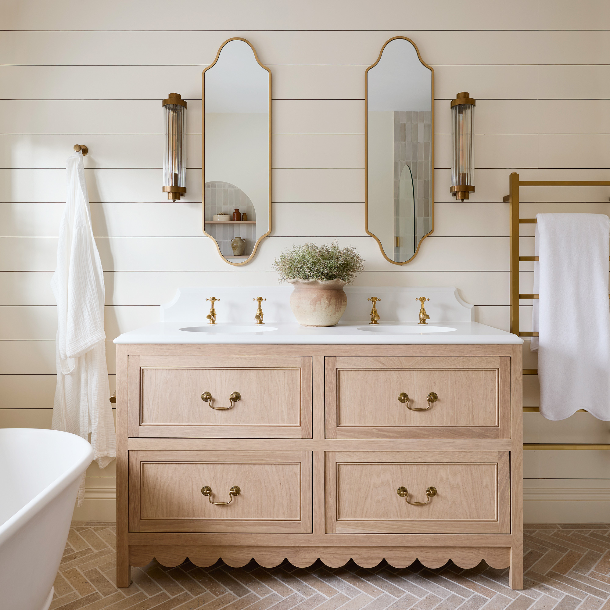 a neutral bathroom with horizontal wall panelling, a large white freestanding bath tub and brass fixtures, double vanity unit, a scalloped edge roman blind and limestone herringbone floor tiles