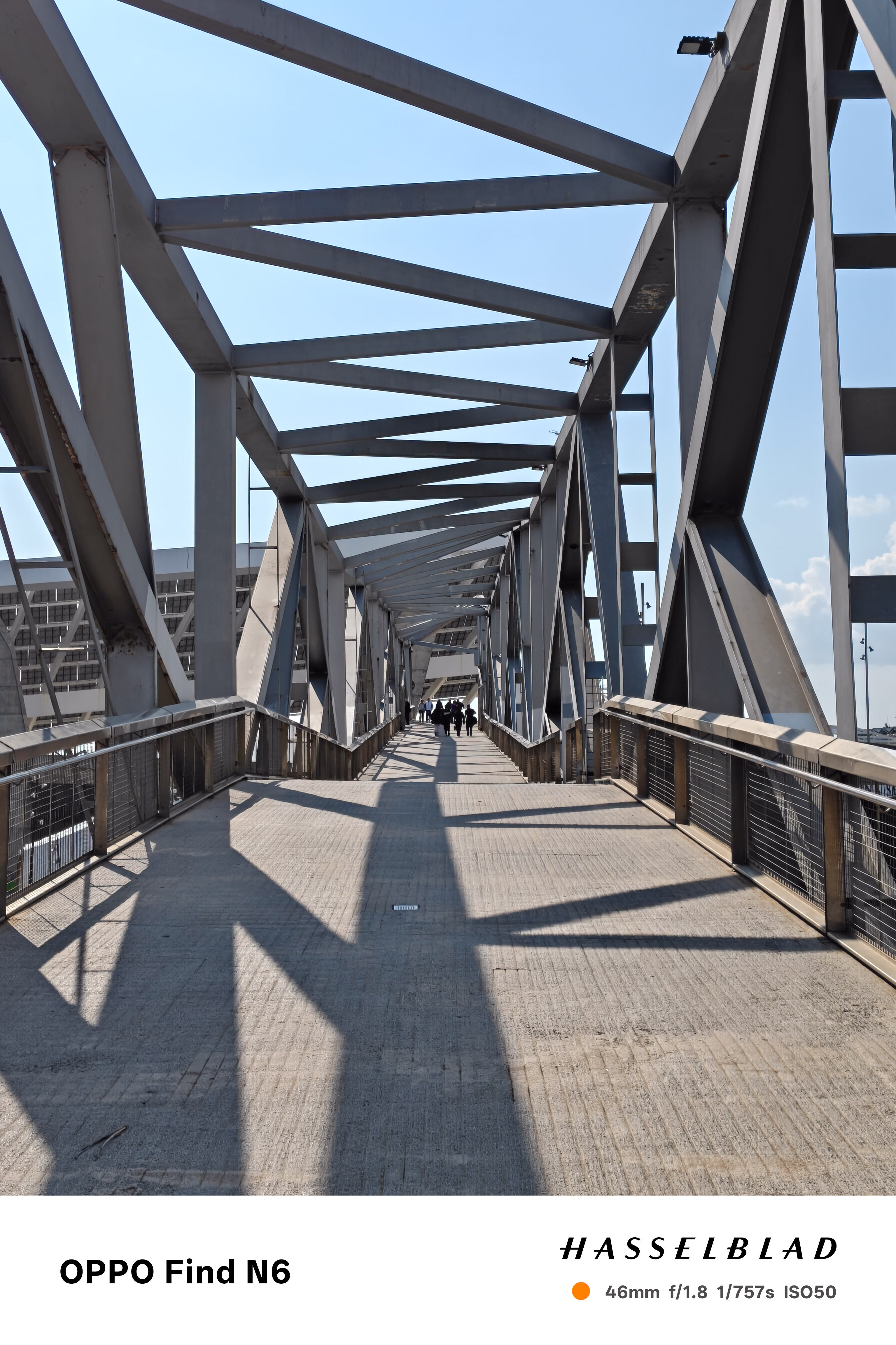 A low-angle perspective shot looking down a long, modern pedestrian bridge with a heavy grey steel truss structure. The sunlight creates dramatic, geometric shadows of the industrial beams across the concrete walkway. A few small figures are seen walking in the distance toward the end of the bridge.