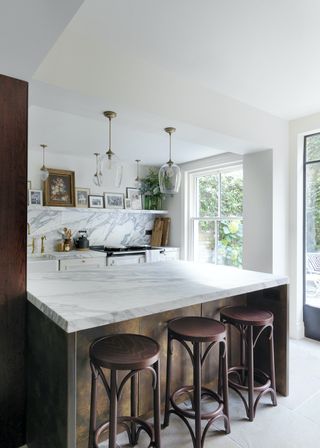 A marble topped peninsula and wooden stools. Above the kitchen islands are two pendant lights. There is also artwork hanging on open shelving.