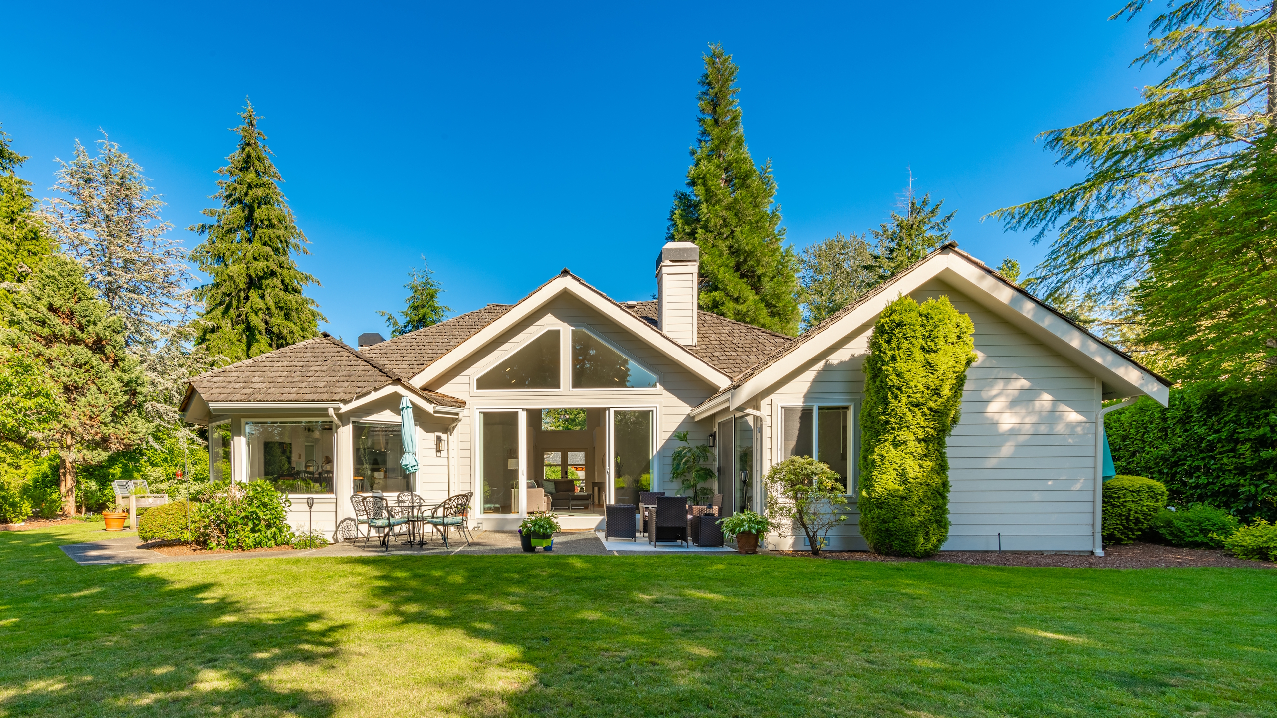 A manicured backyard with trees, shrubs, and a patio seating area around a white house