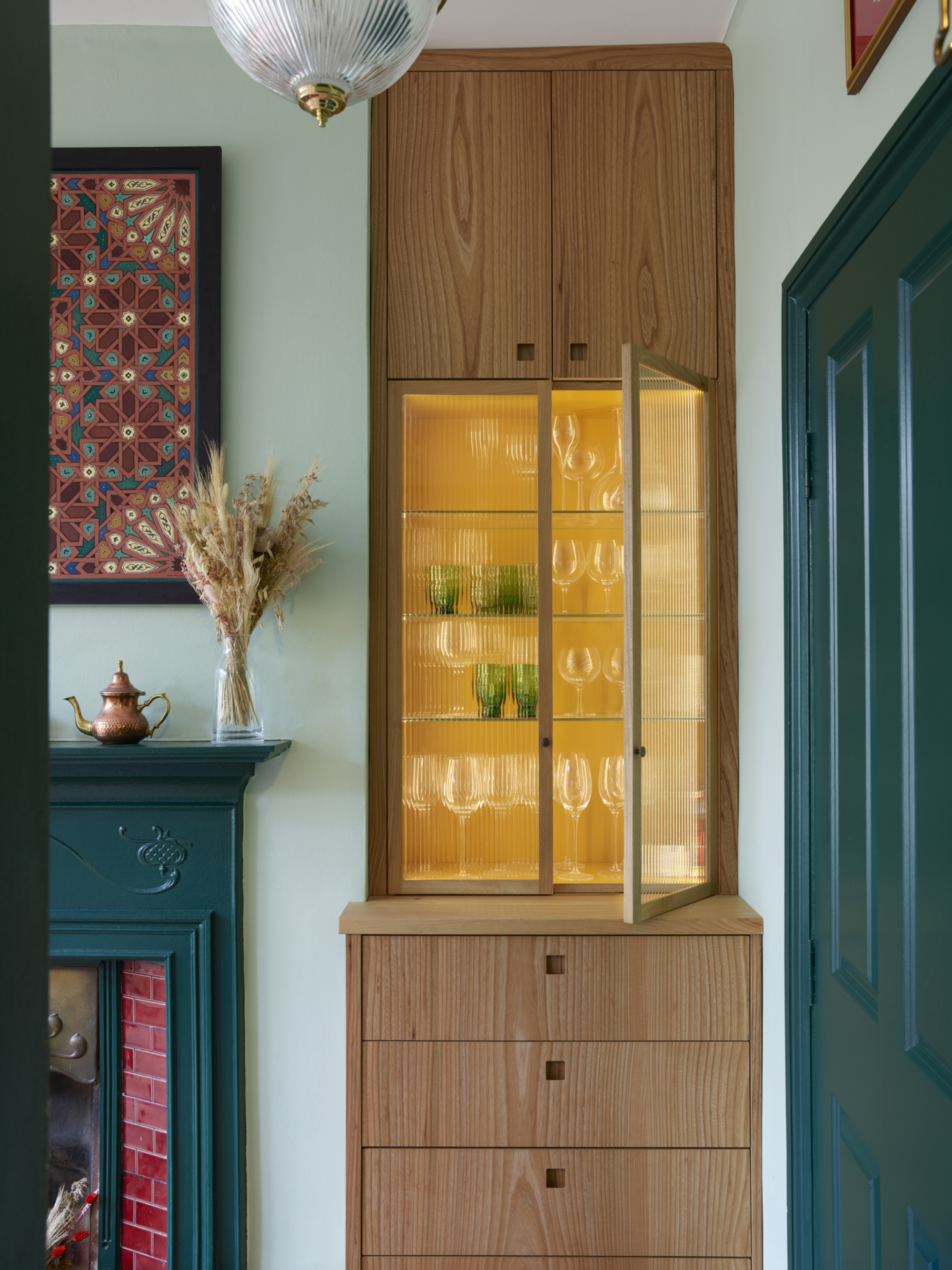 A teal living room with a wooden built-in cabinet and drawers. The built-in has a cabinet portion with glass fluted doors revealing a bright yellow interior.
