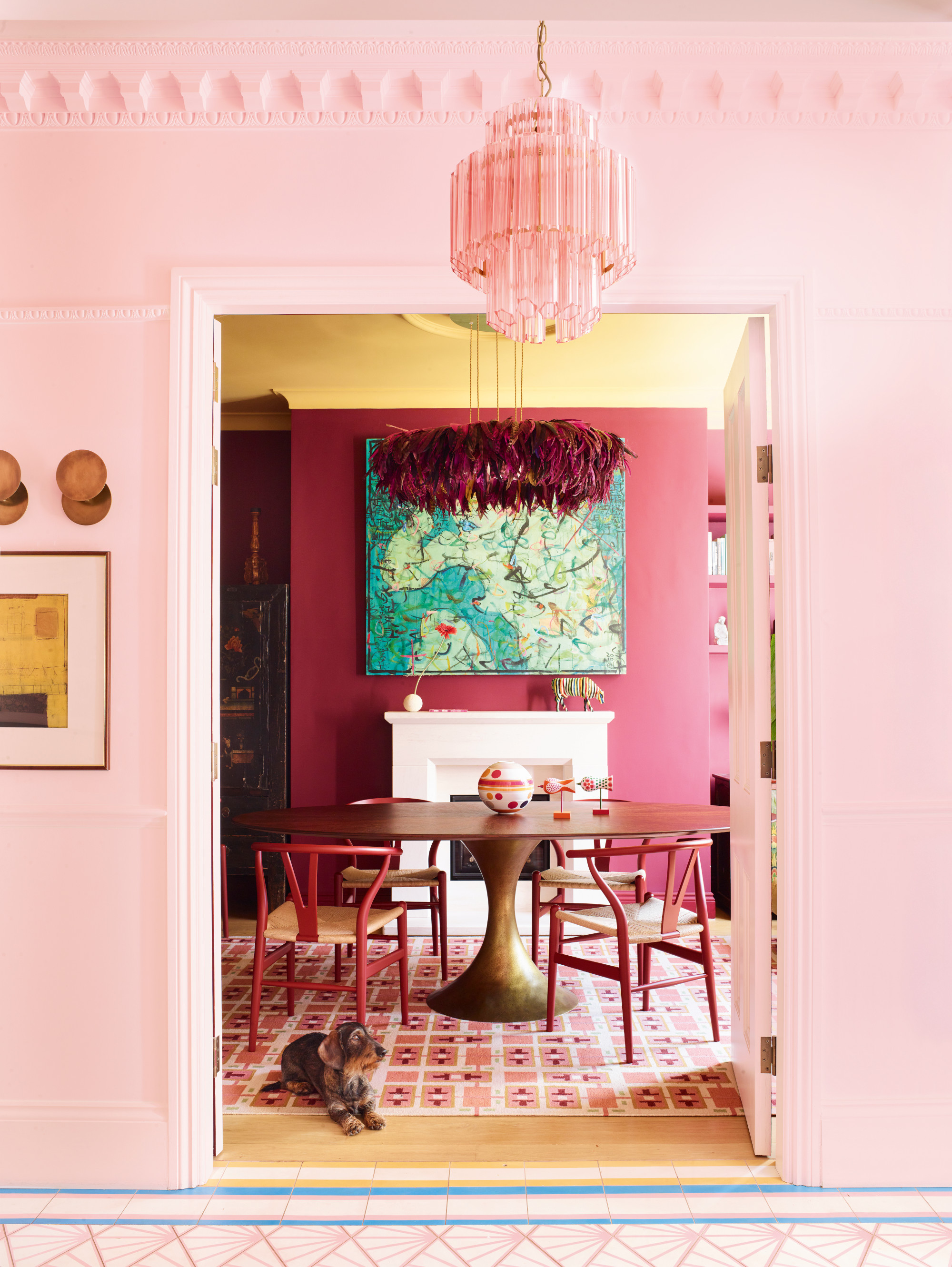 View into dark pink dining room with yellow ceiling, brass pedestal dining table, pink chairs and pink and white patterned rug; green artwork above white fireplace; and burgundy feather chandelier