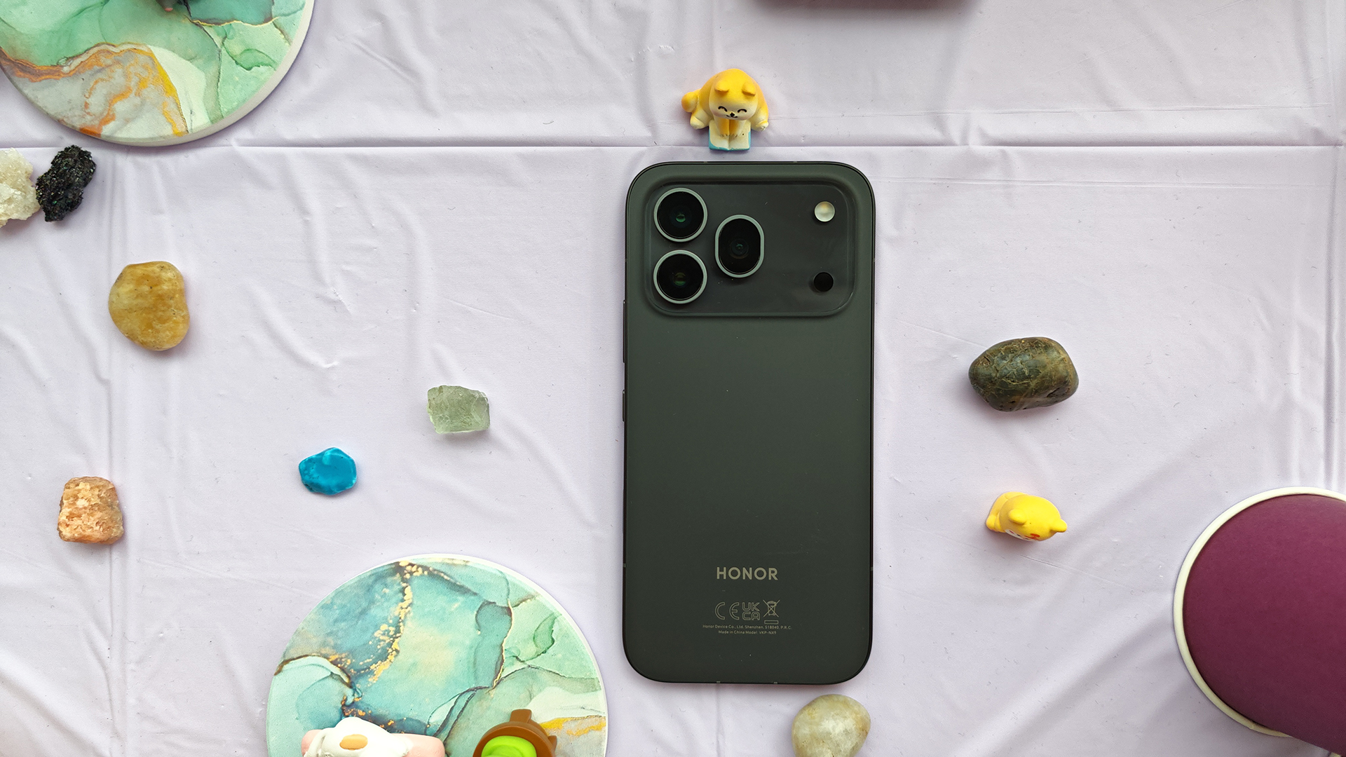 A dark gray Honor 600 Pro smartphone lying flat on a purple surface, surrounded by decorative coasters, stones, and small animal figurines.
