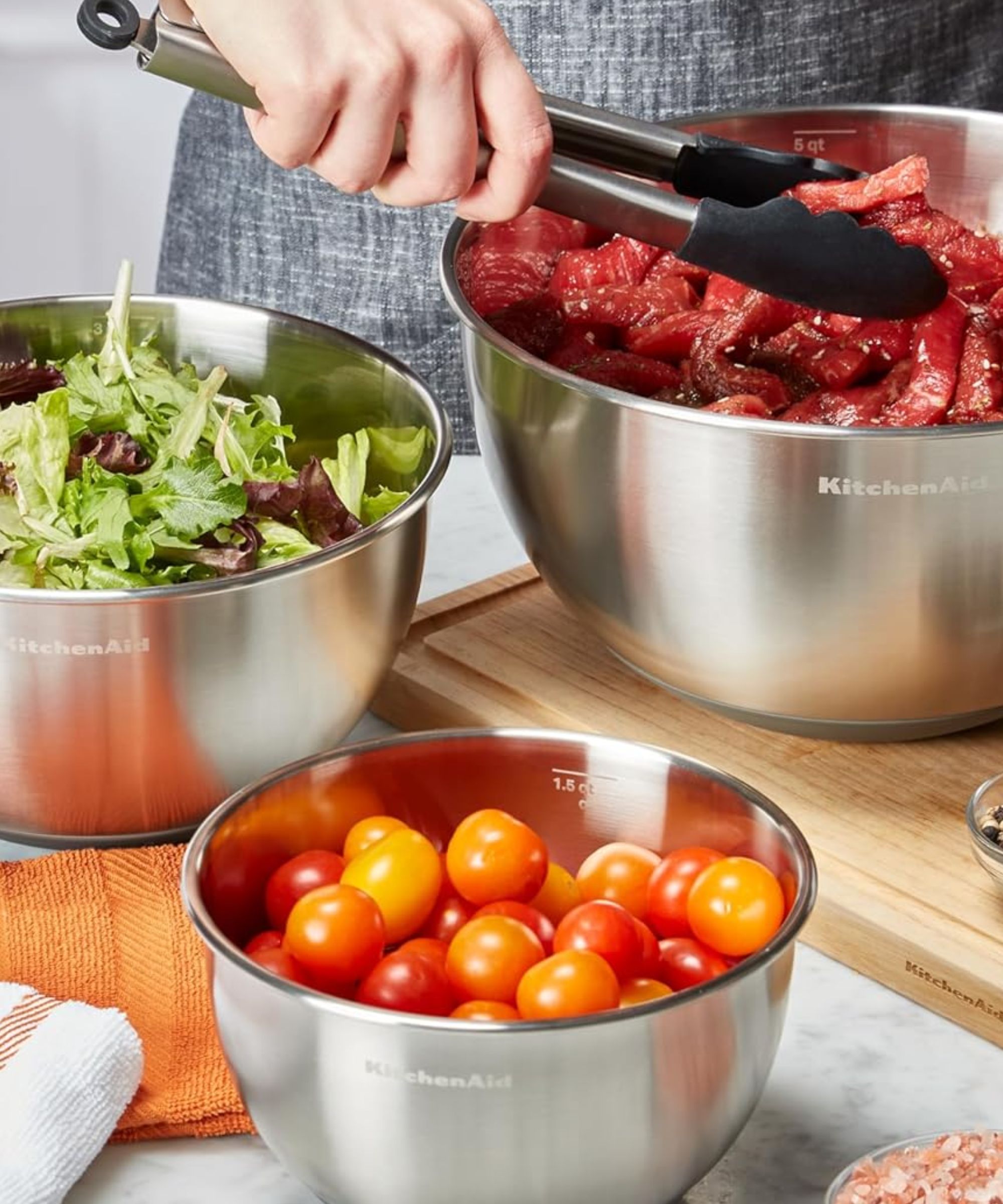 A set of three stainless steel KitchenAid mixing bowls being used in a kitchen being used to prepare meat and vegetables
