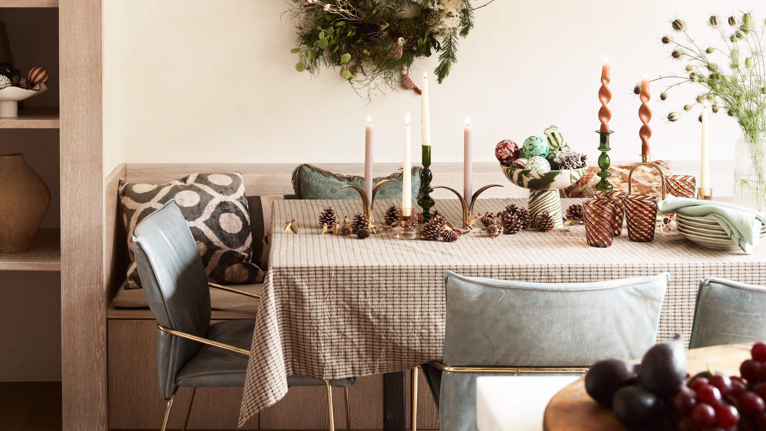 A dining table in a modern kitchen covered with a brown gingham tablecloth and decorated with various dinner candles and a bowl of Christmas baubles