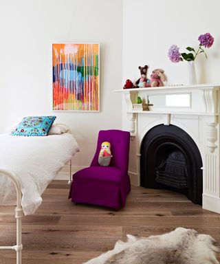 A white child's bedroom with fireplace, wooden flooring, a single cast iron bed, purple chair and animal skin rug.
