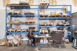 A young woman artist wearing black and brown workwear sits at a desk in a studio filled with raw materials including wood and ceramic while working on a project.