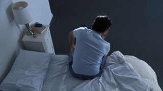A man in a blue t-shirt sits on the edge of a bed because he can't sleep