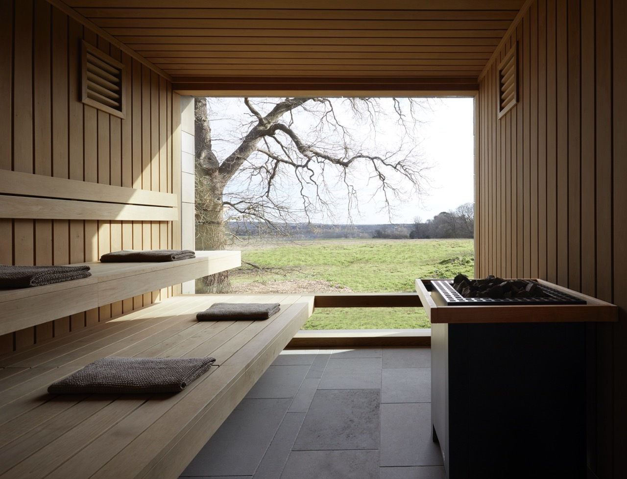 A wooden sauna with tiered benches, each laid with towels, and a wide open window framing a view of a tree and green fields beyond.