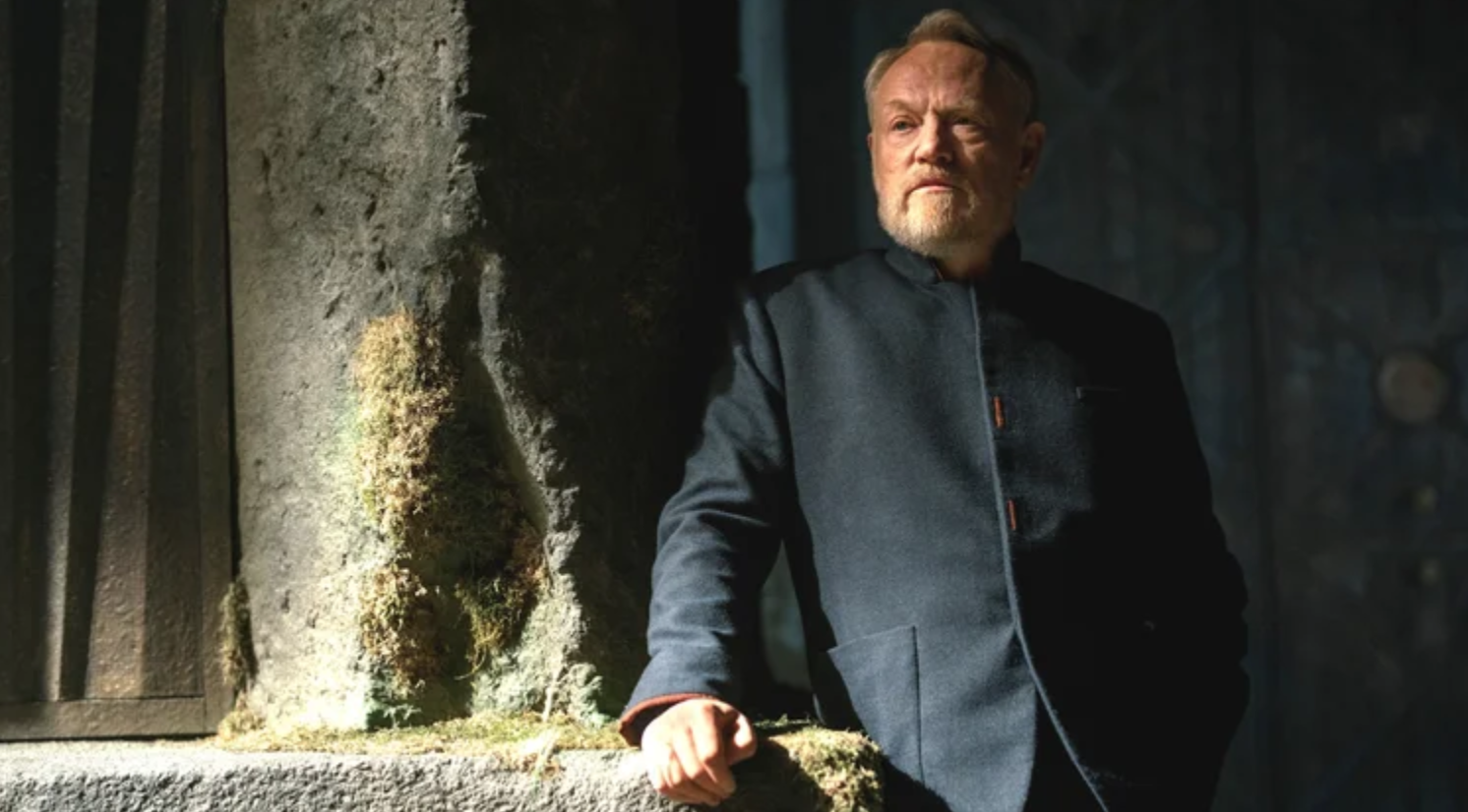 a bearded man stands against a stone pillar