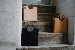3 rectangular shaped shopper style bags with circle handles individually placed on different steps of a concrete staircase. TOP: Beige, MIDDLE: Tan, BOTTOM; Black