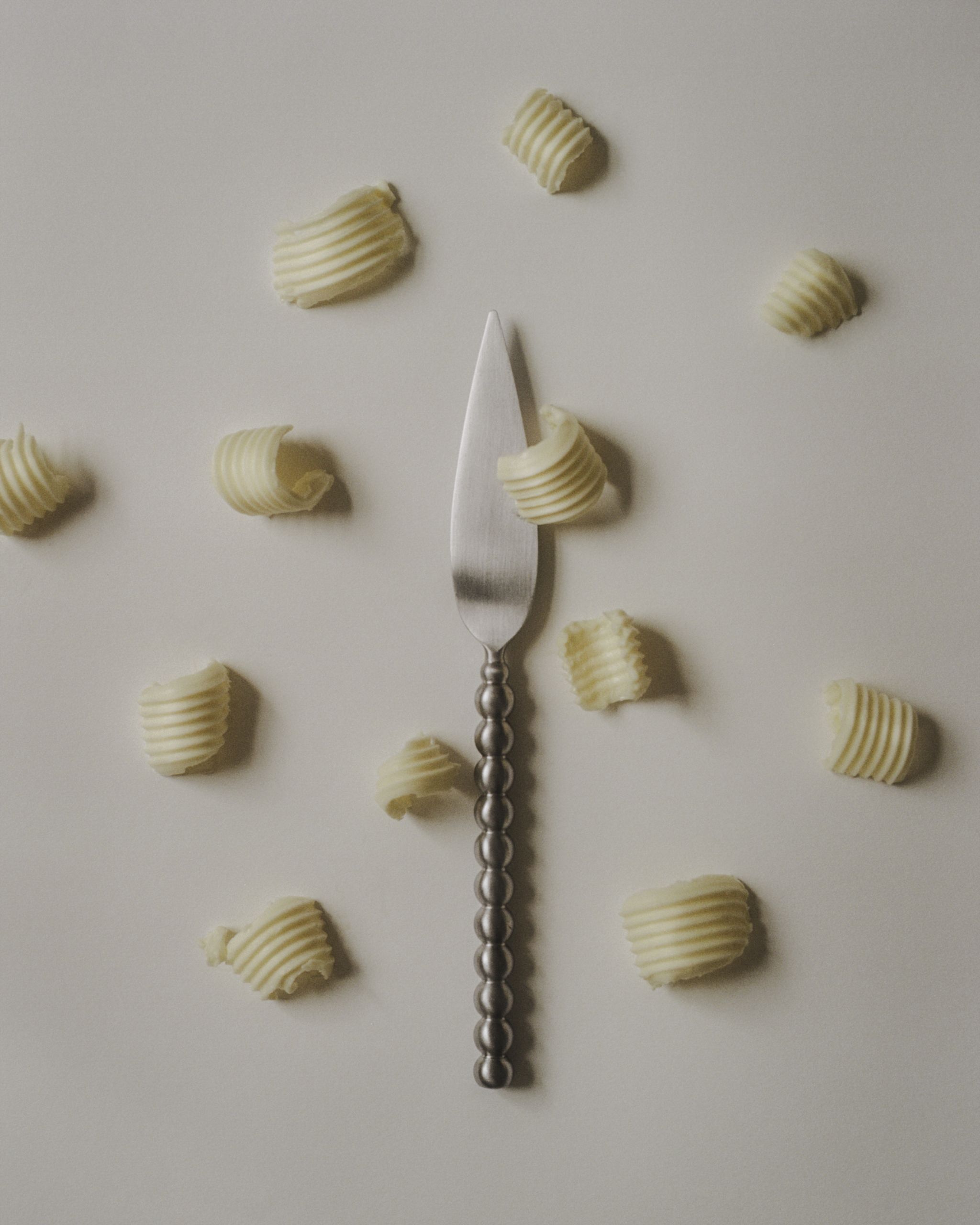 Image of a silver bobbin butter knife on an off-white background with scoops of butter around it.
