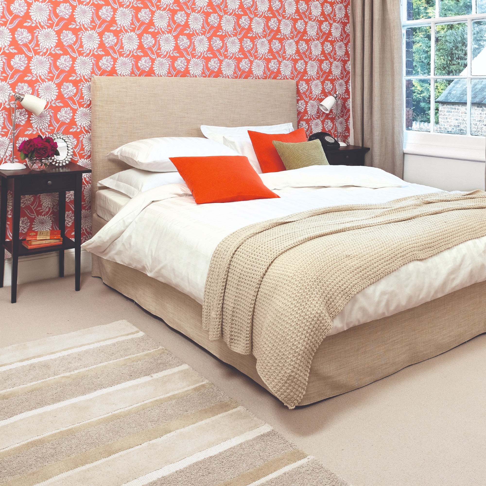 A bedroom in a neutral colour scheme with bright orange cushions and floral wallpaper
