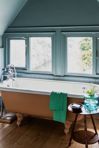 blue bathroom with painted shiplap, rust coloured painted tub, wooden floorboards and side table