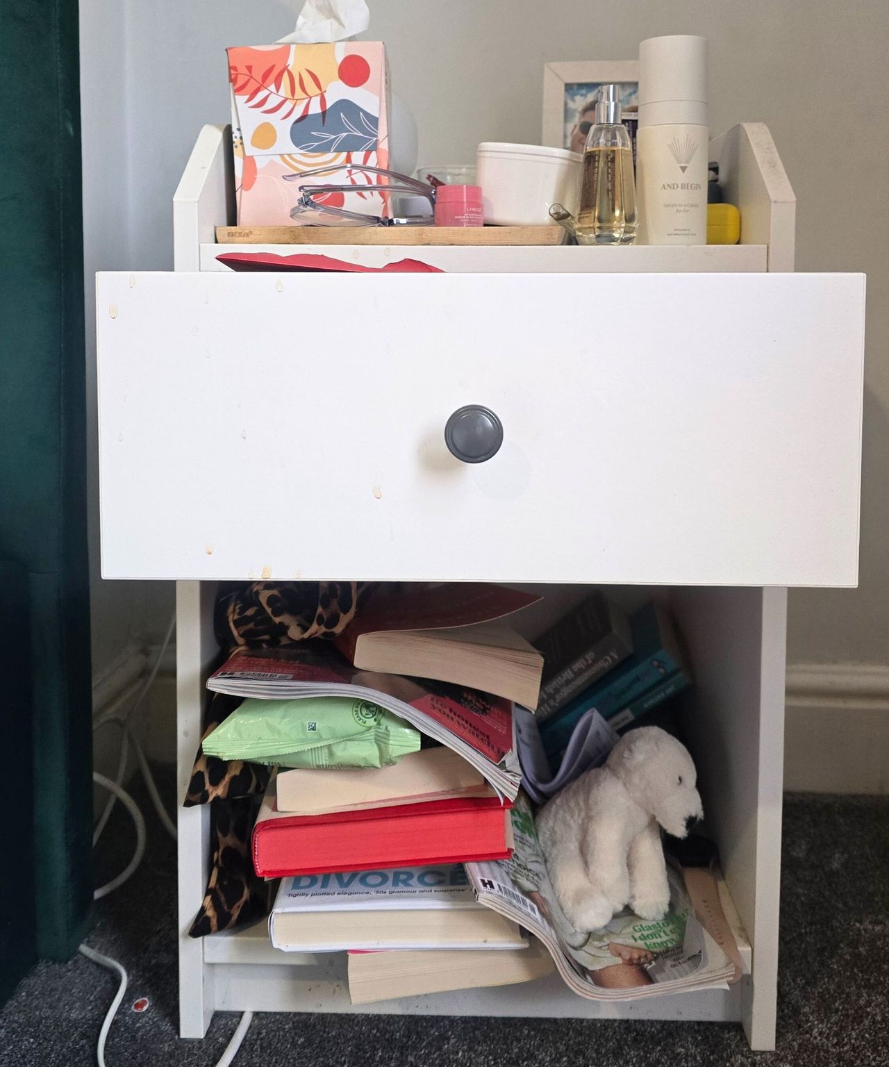 A white nightstand with colorful books and a polar bear piled up on the bottom shelf, an open drawer, and a top shelf with perfume, skincare bottles, and a box of tissues on it