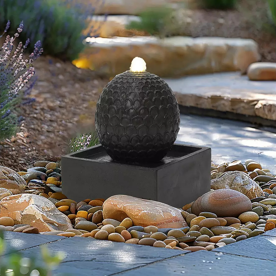 water feature with square base, round ball and light at top in garden with large stone-filled borders