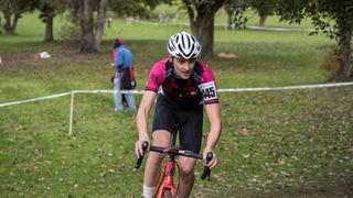 A white man in a pink and black jersey rounds a corner on a grass track