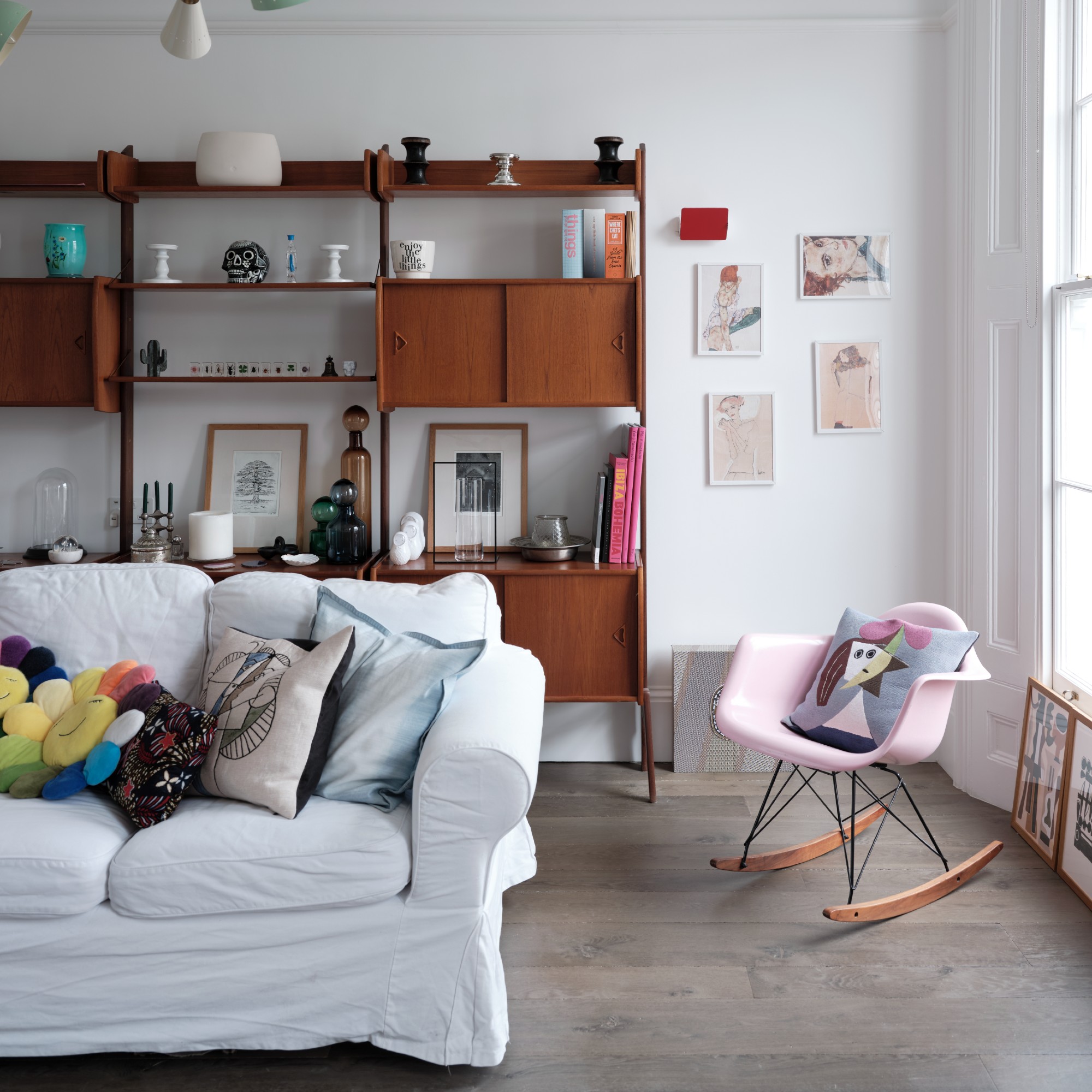 A white living room with a white sofa with colourful scatter cushions, a pink rocking chair and a mid-century modern bookcase