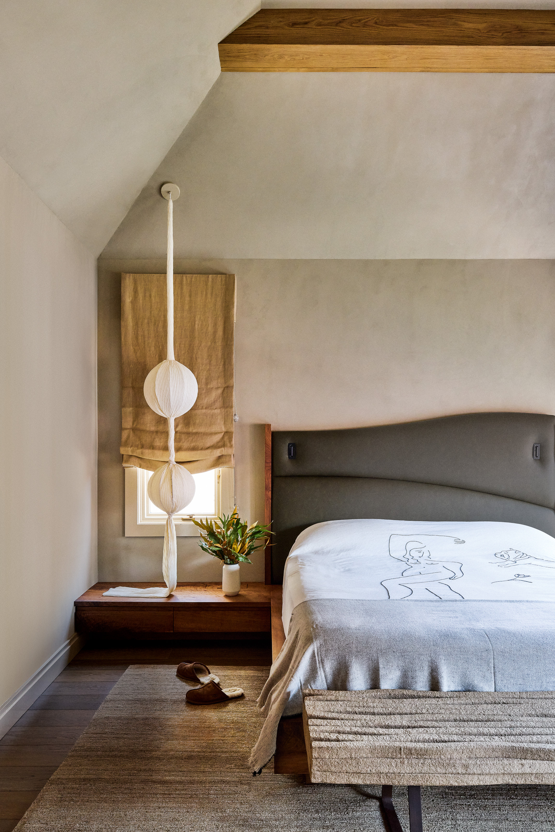 Bedroom with beige plaster walls, grey curved headboard, low bedside table and upholstered grey fabric bench