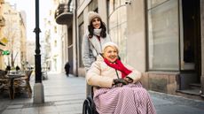 A daughter or volunteer pushes an older woman (or her mother) in a wheelchair on a city sidewalk. They appear happy to be out and about.