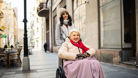 A daughter or volunteer pushes an older woman (or her mother) in a wheelchair on a city sidewalk. They appear happy to be out and about.